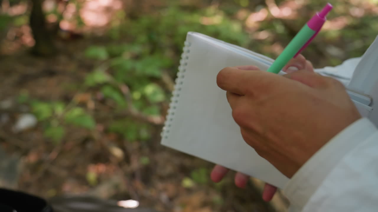 Close view of hand placing pen on jotter while standing outdoors, showing focus on writing and documentation with blurred natural background creating calm forest atmosphere suitable for research