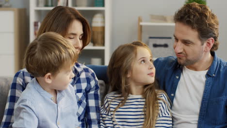 cerca de los padres jóvenes sentados en el sofá de la sala de estar y hablando con su pequeña hija linda y su hijo que se sientan de rodillas