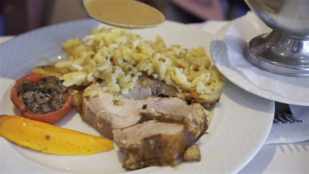 Close up of a server arranging a portion of egg noodle near a few pieces of pork, cooked carrot and stuffed tomato on a white plate