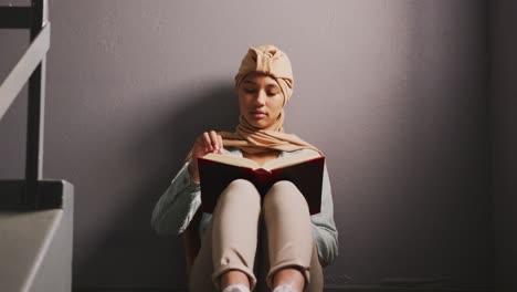 Asian-female-student-wearing-a-beige-hijab-sitting-on-a-staircase-and-reading-a-book