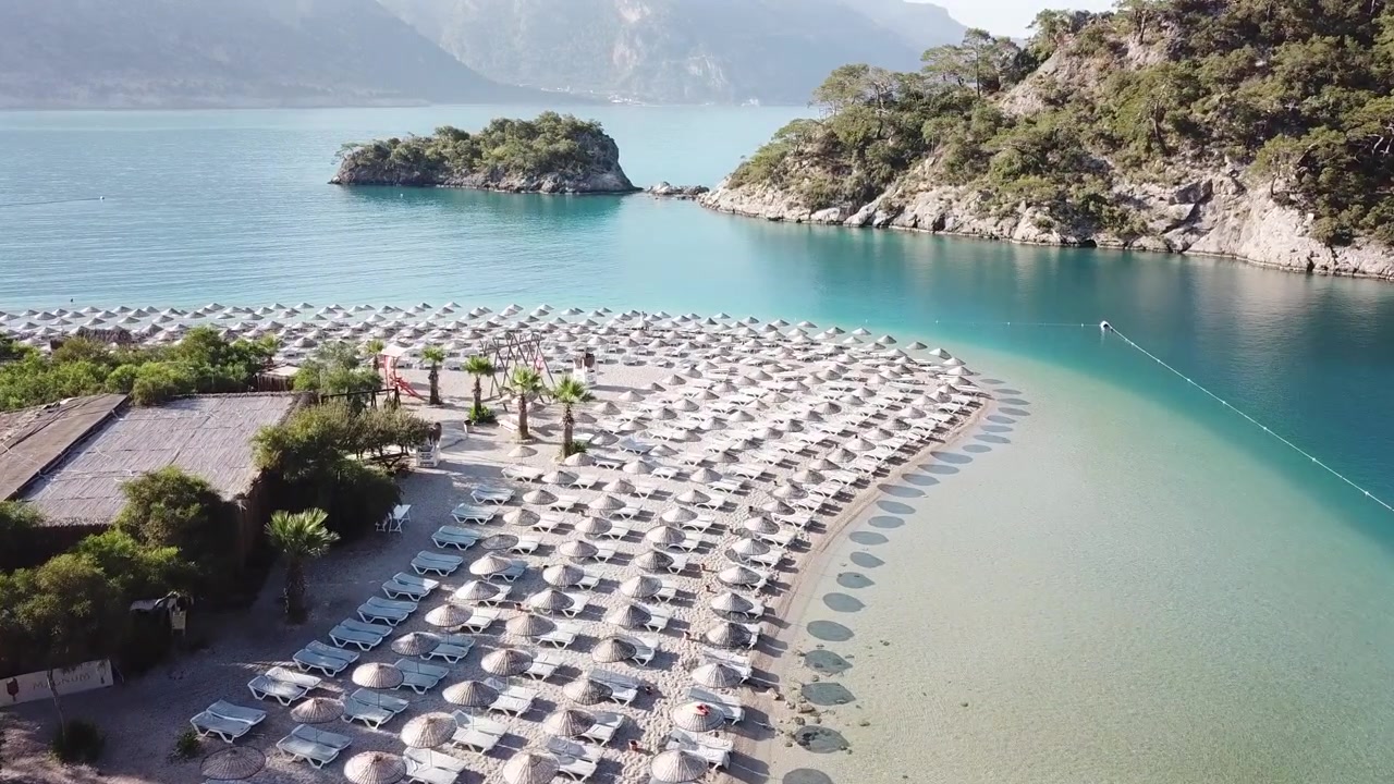 Turkki, Fethiye, Ölüdeniz (Kuollutmeri). Droonikuvaa aurinkoisena päivänä turkoosin laguunin yllä kohti niemeä, jossa näkyy aurinkotuoleja ja aurinkovarjoja varjoineen. Rauhallinen maisema.