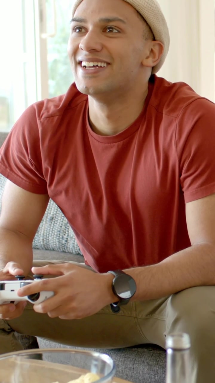 Vertical video: Reacting to game event, man in red tee pressing white controller on sofa with bowl