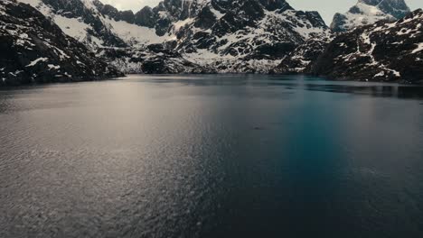 flying over the calm water of lake to the rocky mountain with snow in winter in lofoten island, norway. - aerial shot