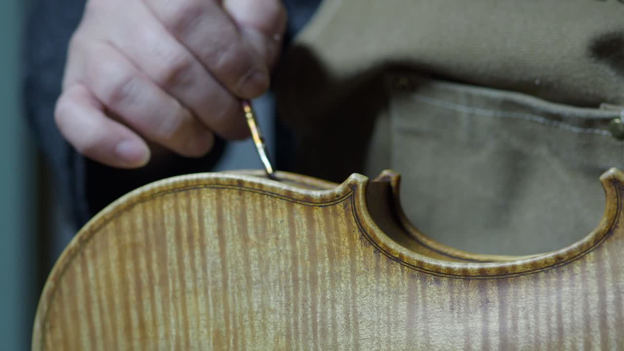 Static close-up of luthier antiquing new violin, rubbing glaze with fingers on the violin’s rib, revealing detailed marbling and natural wood flames pattern on back plate in traditional workshop