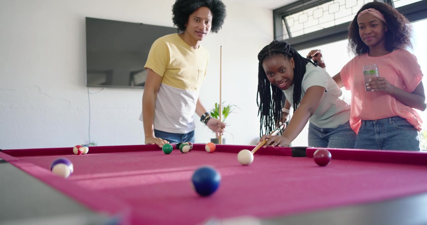 African American friends at home laughing, woman striking cue ball on red-felt table to take shot