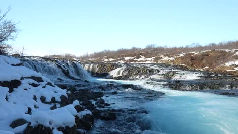 wide ground-level view of brúarfoss waterfall, iceland. vivid turquoise water cascades over dark rocks in a partially snow-covered winter scene.