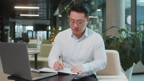 asian businessman writing down notes while attending online video class in modern office lobby