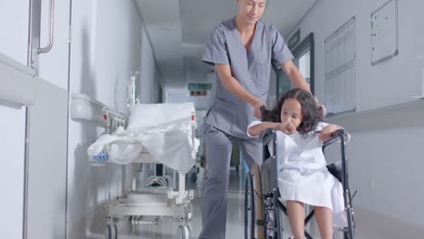 Diverse-male-nurse-and-child-patient-sitting-in-wheelchair-in-corridor-at-hospital,-in-slow-motion