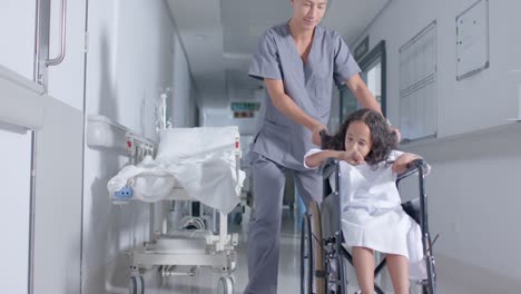 diverse male nurse and child patient sitting in wheelchair in corridor at hospital, in slow motion