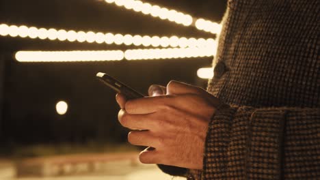 young man texting chatting on smart phone at night; men finger tapping keypad, writing message on mobile devices touchscreen, blue low light; extreme close up top view