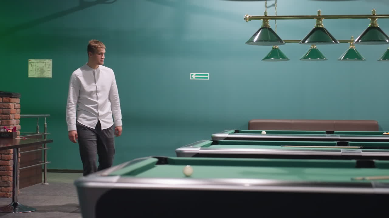 Uomo in camicia bianca e pantaloni grigi che entra in una sala da biliardo circondato da tavoli da biliardo verdi e lampade a sospensione. Espressione concentrata, atmosfera elegante da club sportivo, ambiente ricreativo moderno