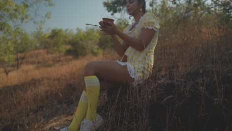 panorámica en cámara lenta hacia abajo de una atractiva joven india vestida de amarillo sentada en una roca, sosteniendo cerámica en el campo