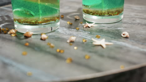 decorating materials including small beads, star-shaped and shell-like items are scattered on kitchen work table beside tealight cups filled with green and golden wax mixture