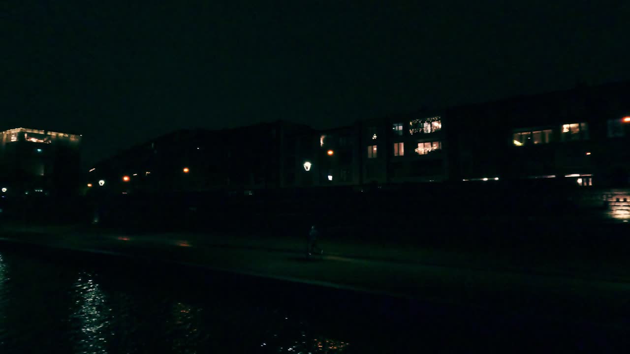 Persone camminano lungo una strada mentre gli edifici con le luci brillano nell'oscurità. La location presenta un mix di scene residenziali e commerciali. Un fiume riflette le luci.
