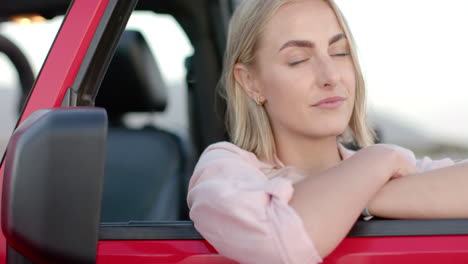 young caucasian woman with blonde hair smiles gently, leaning on a red car door during a road trip