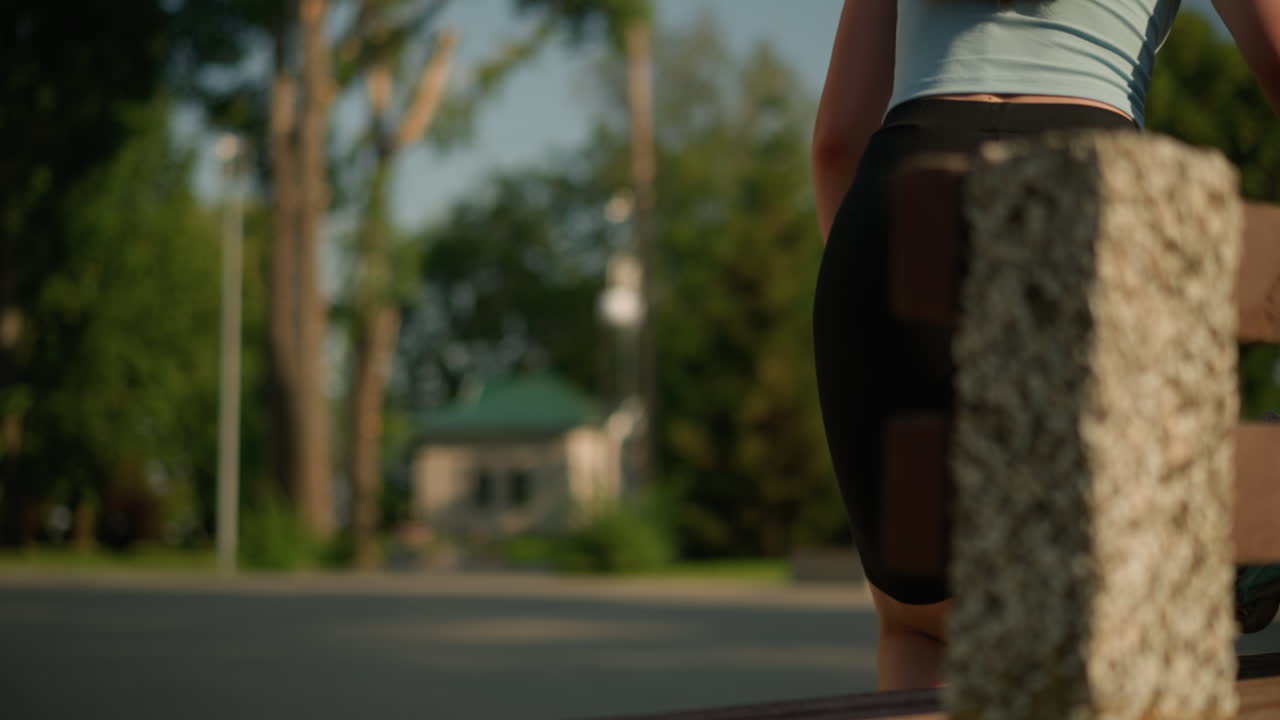 시안색 롤러 스케이트를 들고 나무 공원 벤치에 떨어뜨리는 젊은 여성, 그녀는 검은색 양말과 밝은 파란색 을 입고 높은 나무, 초록색, 그리고 램프 포스트를 특징으로하는 흐릿한 배경