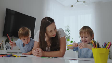 A-young-mother-with-two-children-sitting-at-a-white-table-draws-colored-pencils-on-paper-in-slow-motion