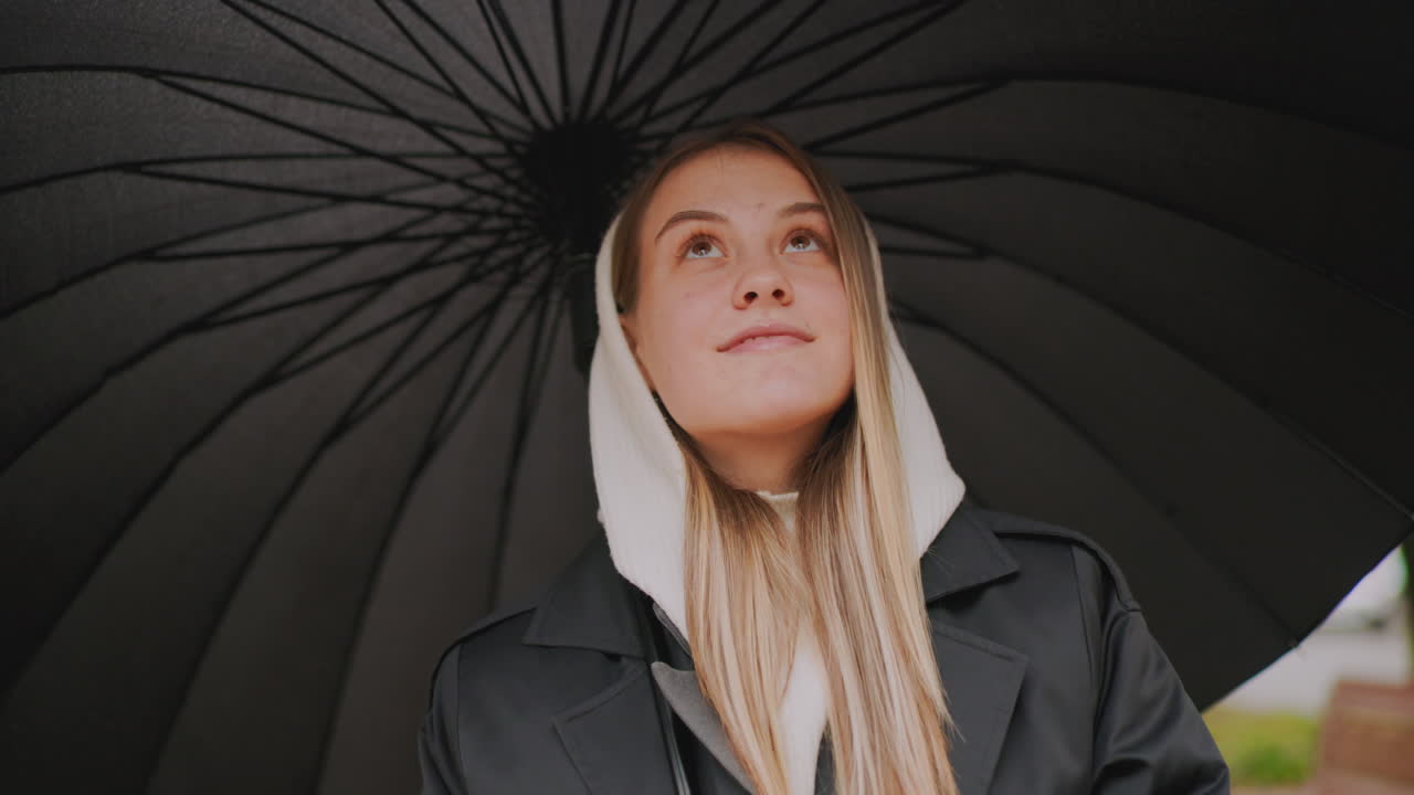 Young woman with blonde hair in hooded jacket standing under umbrella during rainy day, looking sideways with gentle smile, holding hair strand, natural candid moment outdoors in overcast weather atmosphere
