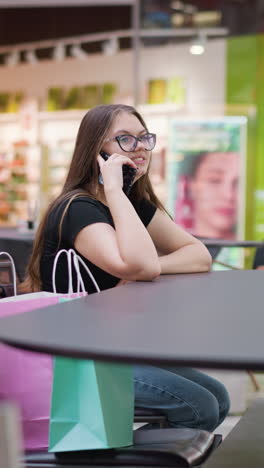 joven elegante con gafas felizmente hablando por teléfono, sonriendo, mirando hacia la derecha, sentada en un moderno patio de comidas con fondo borroso con estructuras y mesas, con el transeúnte visible en la distancia