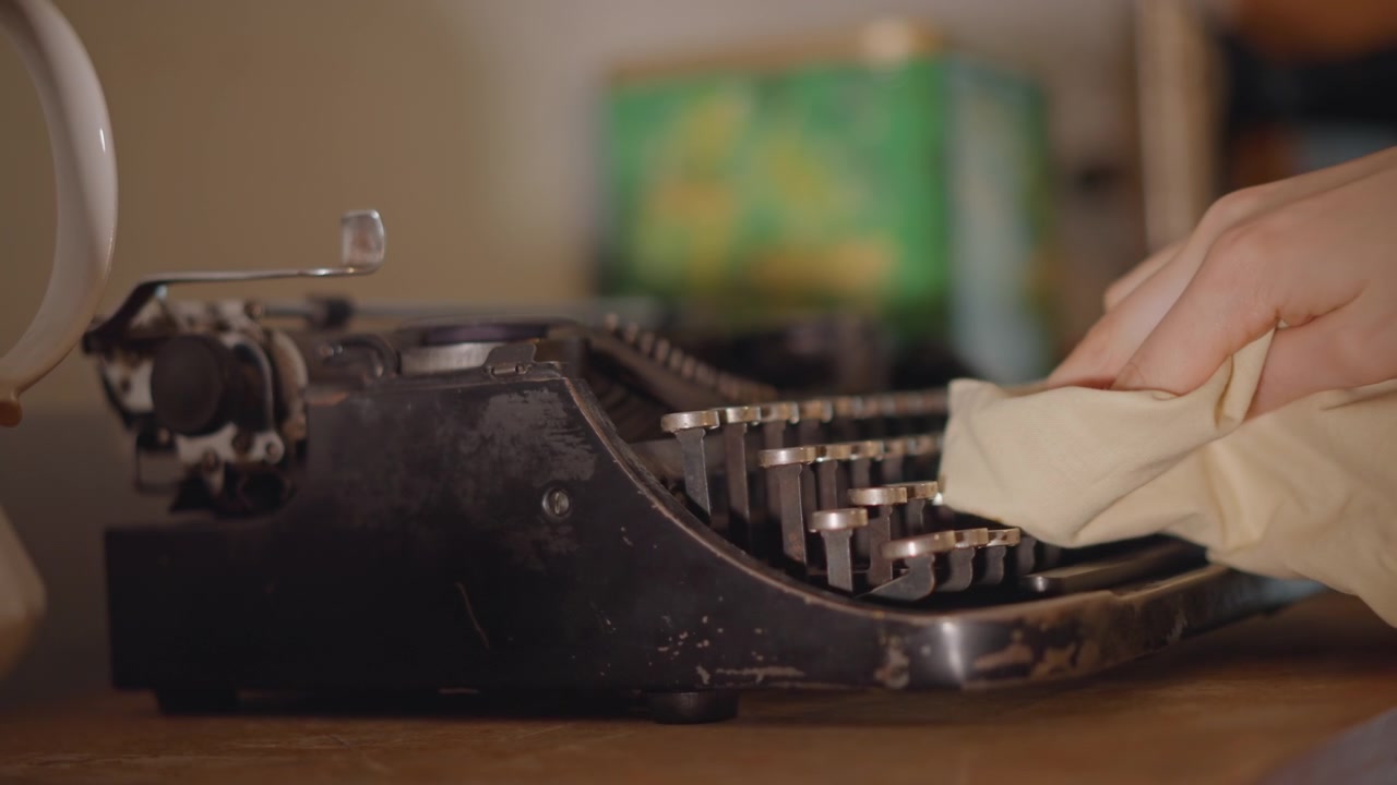 Primo piano di tasti di macchina da scrivere vintage in fase di pulizia, mani attente che rimuovono la polvere dai tasti metallici in un accogliente laboratorio, calda illuminazione cinematografica che crea un'atmosfera nostalgica, rullo testurizzato e alloggiamento del nastro visibili.