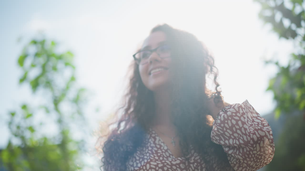 Woman with glasses and cross pendant flaunts her hair outdoors, smiling warmly as sunlight reflects off her face, surrounded by lush greenery and gentle natural light