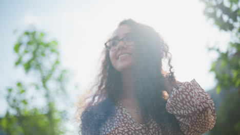 mujer con gafas y colgante de cruz exhibe su cabello al aire libre, sonriendo cálidamente mientras la luz del sol se refleja en su cara, rodeada de exuberante vegetación y suave luz natural