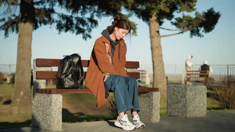 a girl sits alone on a bench in an urban park, looking deeply melancholy with her head resting on her hand. she is wearing a brown coat, blue jeans, and white shoes, with a black backpack beside her