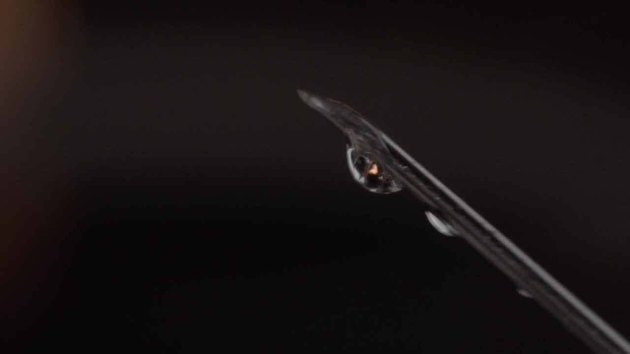 Macro primo piano di un ago di siringa con una goccia di medicinale che si forma sulla punta affilata su sfondo scuro, evidenziando l'attrezzatura medica precisa e il dettaglio del liquido