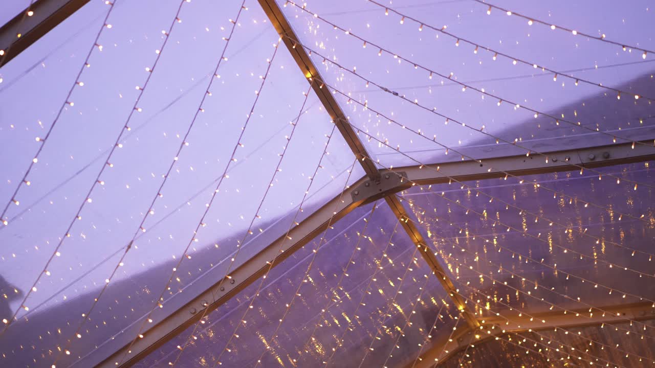Strings of warm fairy lights suspended on a transparent roof structure during dusk