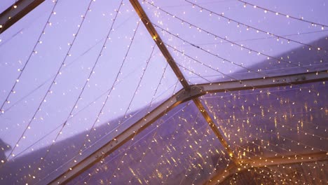 strings of warm fairy lights suspended on a transparent roof structure during dusk