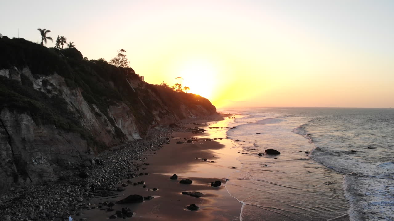A sand beach at sunrise with ocean waves and colorful water reflections on the shore with cliffs in silhouette on the Santa Barbara, California coast AERIAL DRONE