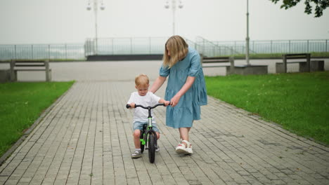 a mother is gently guiding her young son as he rides his bicycle along an interlocked pathway, the boy appears focused, while his mother helps to steady him