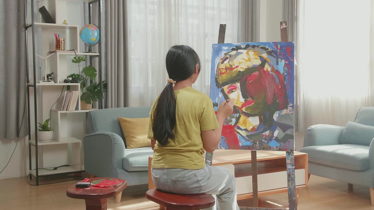 Hind View Of Asian Artist Girl Sitting On Round Wooden Chair Without Backrest Holding Paintbrush Mixed Colour And Thinking Before Painting A Girl'S On The Canvas