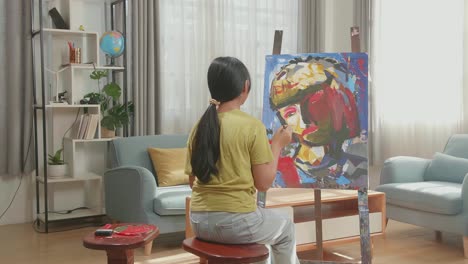 hind view of asian artist girl sitting on round wooden chair without backrest holding paintbrush mixed colour and thinking before painting a girl's on the canvas