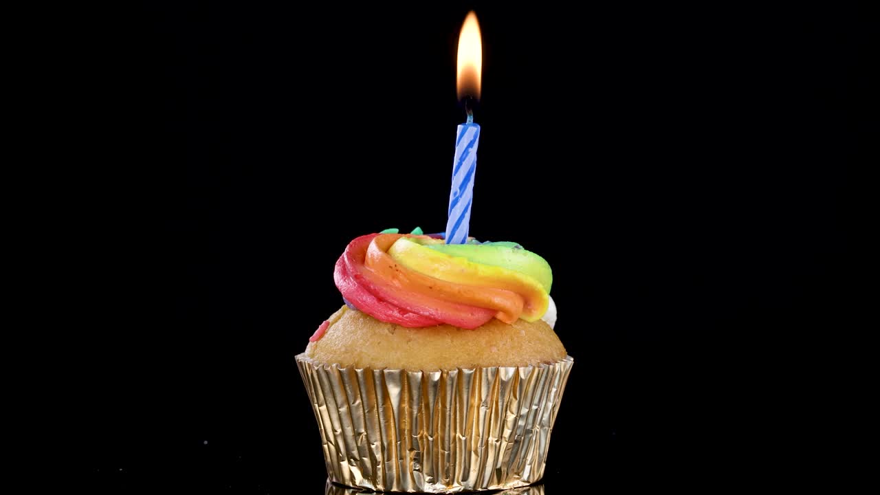 A single cupcake with rainbow frosting and a blue-striped lit candle stands against a black background. Steady camera, dramatic lighting, celebratory mood