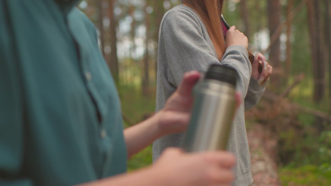 Close-up of young woman in green shirt pressing inner cover of thermos to access water in peaceful forest, while another woman, slightly blurred in background, brushes her hair
