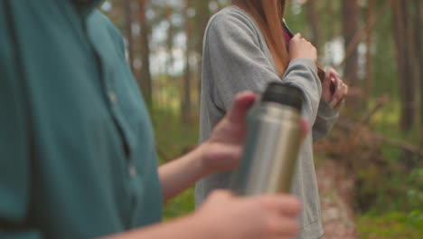 close-up of young woman in green shirt pressing inner cover of thermos to access water in peaceful forest, while another woman, slightly blurred in background, brushes her hair