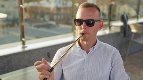 mid aged man wearing sunglasses and shirt sits outdoors. male resting and smoking hookah producing a cloud of thick white smoke.