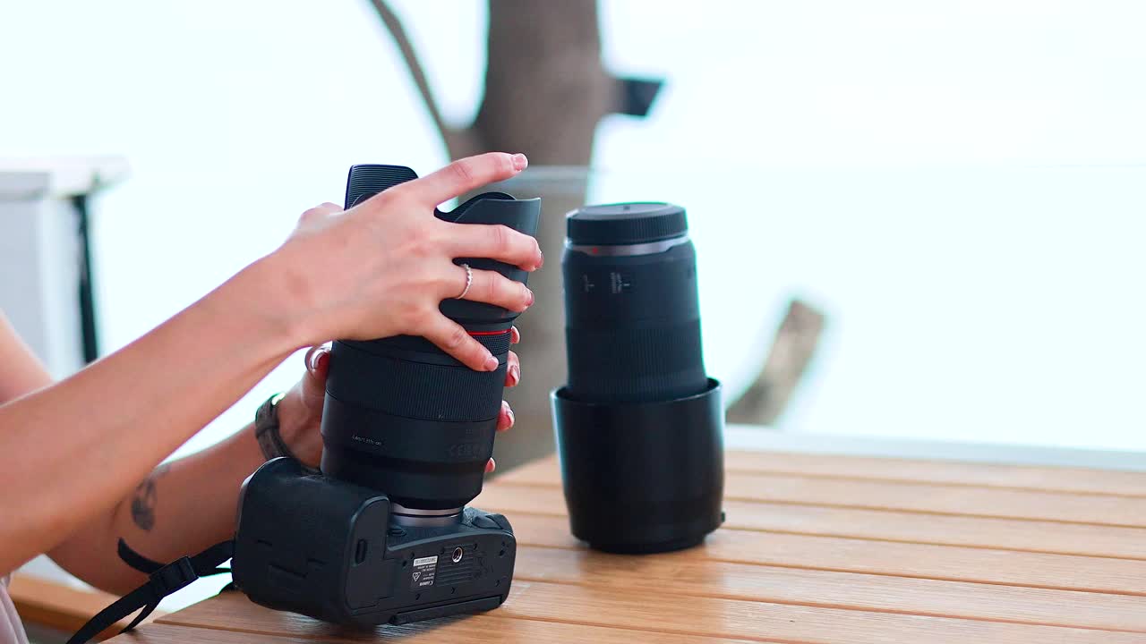 A woman attaches a lens to a camera on a wooden table. Bright outdoor lighting enhances the serene Phuket setting