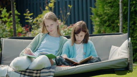 two cute girls read books, sit on a garden swing in the backyard of the house