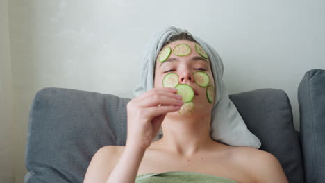 lady gently removes cucumber slice from eyes with hand, about to chew it while resting with cucumber mask and towel wrap in calm setting, enjoying refreshing skincare, wellness