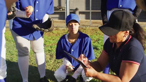 diverse group of female baseball players listening to female coach, squatting and talking on pitch