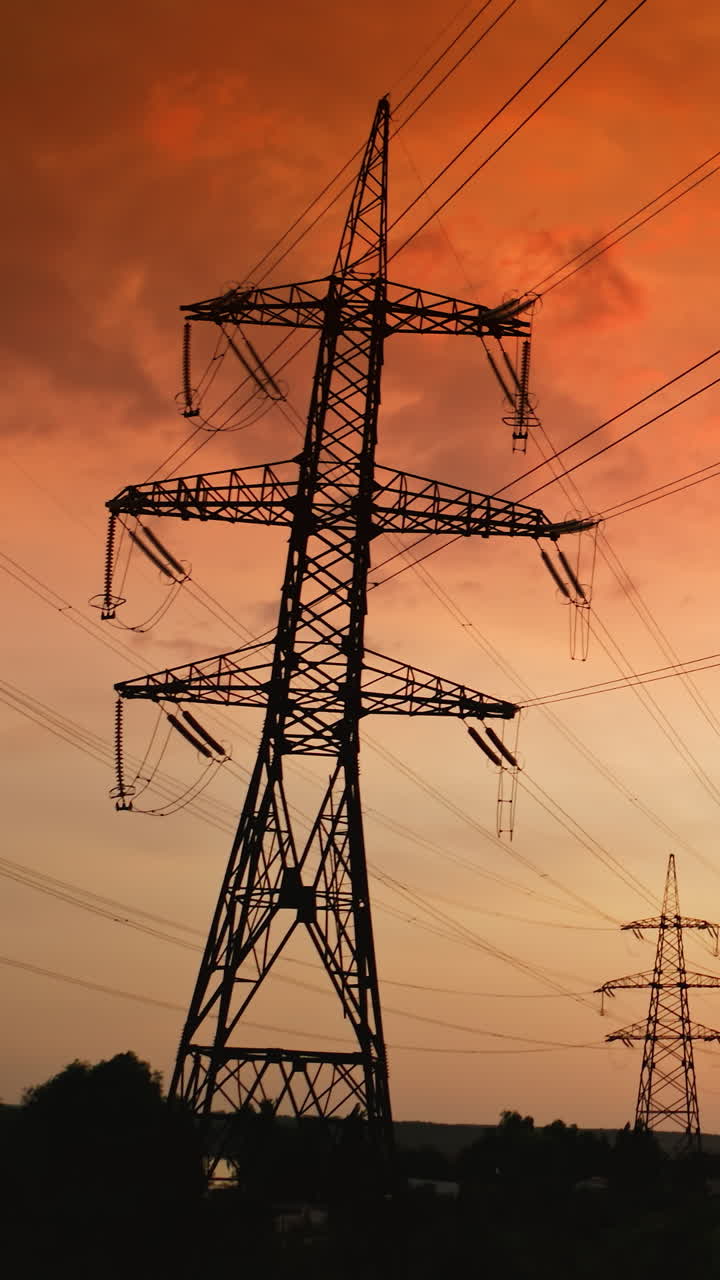 Transmission lines at dusk. Silhouette of high voltage electric supports against red sky. Dark smoke from industrial pipe going into the air near electrical pylons. Vertical video