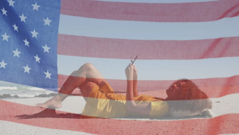 animation of american flag waving over woman using tablet lying on beach