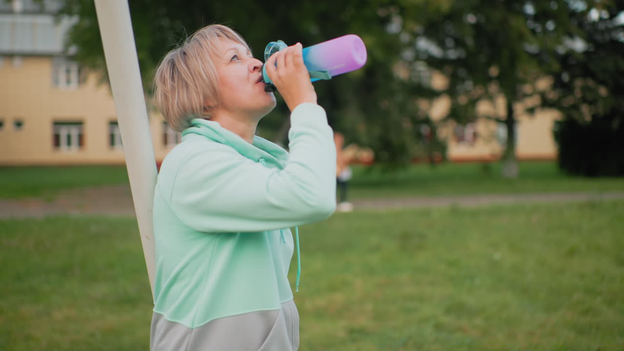 Instructor pausing during outdoor activity to drink water from colorful bottle while distant figure moves through garden background under bright sky capturing peaceful break moment relaxation