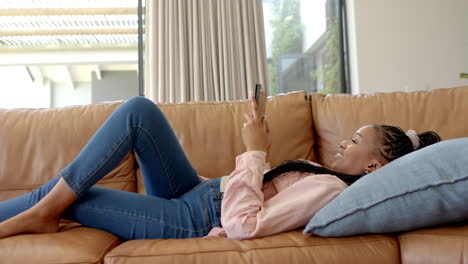 african american young woman lying on couch, holding smartphone