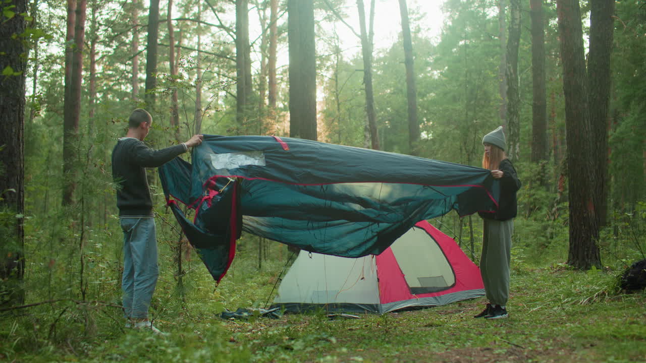 Young couple working together to spread out and arrange tent fabric on forest ground during outdoor camping, surrounded by tall trees and natural greenery with pitched tent visible in background