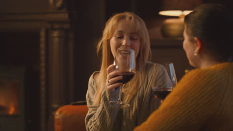 two female friends relaxing and making toast on sofa in lounge with cosy fire holding glass of wine