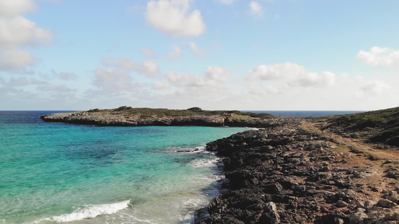 aerial drone shot of a rocky coastline and a red dirt path with waves from the brilliant blue water of the Mediterranean crashing onto the rocks of Majorca