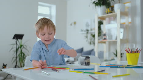 Lächelnder-Junge-Im-Blauen-Hemd-Zeichnet-Mit-Einem-Bleistift-Auf-Papier,-Während-Er-Am-Tisch-Im-Wohnzimmer-Sitzt